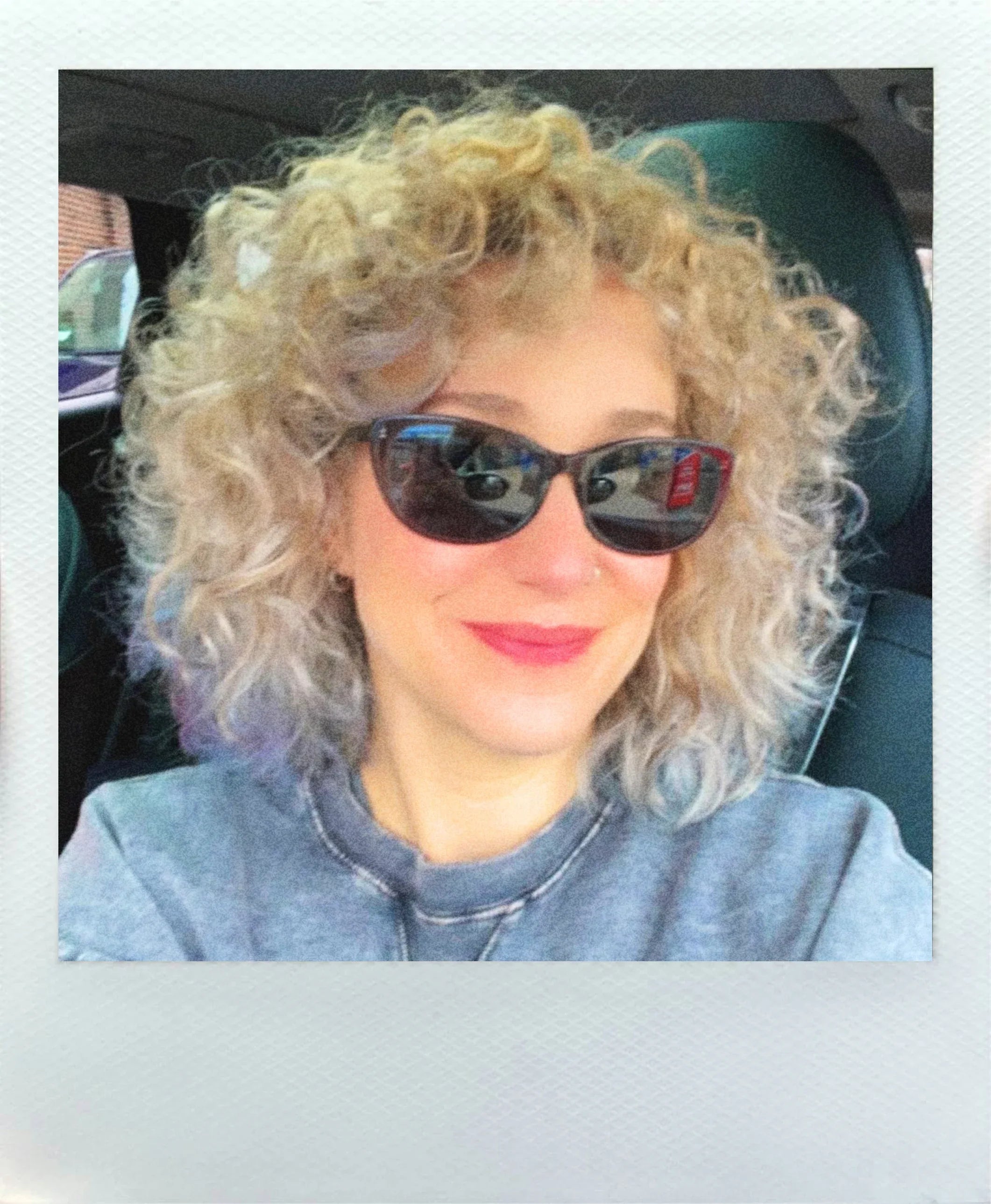 Smiling person with curly blonde hair and sunglasses sitting in a car, wearing a grey top.