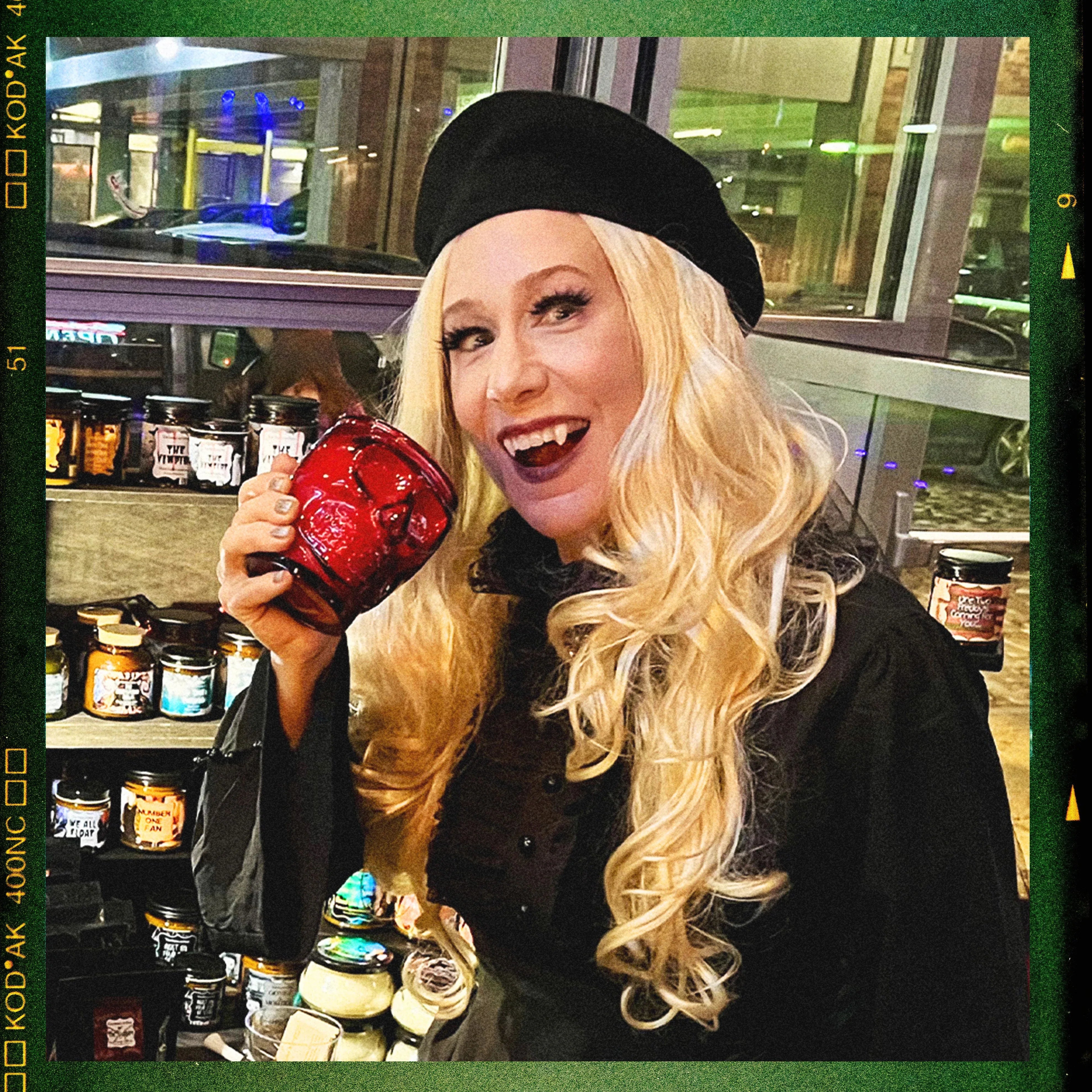 Woman in gothic attire holding red horror-themed candle, surrounded by spooky candles display