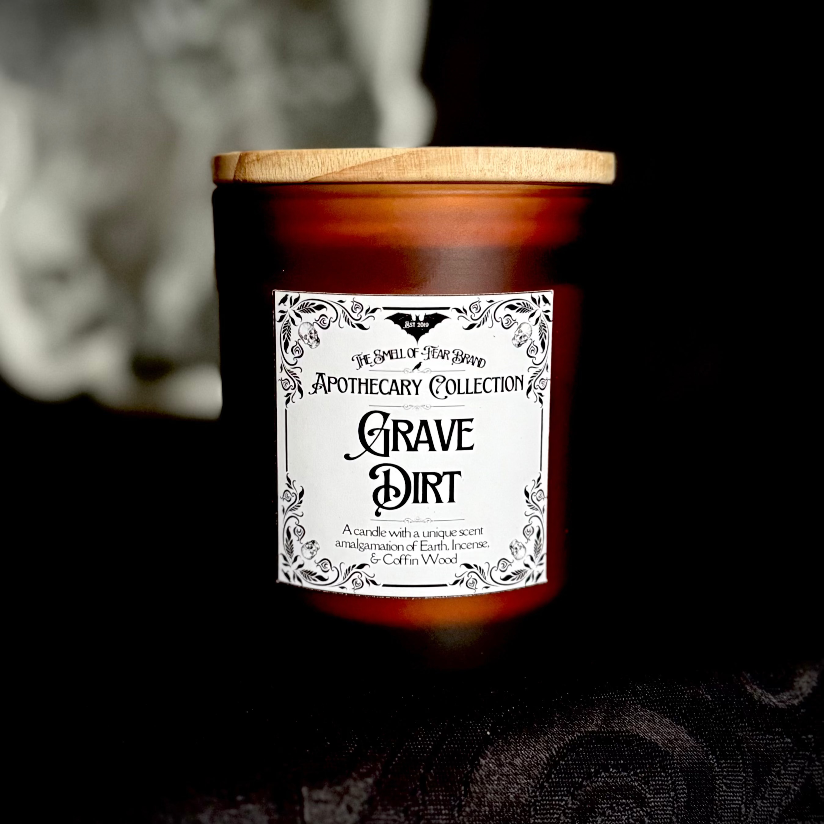 A candle labeled 'Apothecary Collection: Grave Dirt' with a wooden lid, set against a backdrop featuring trees and a full moon.