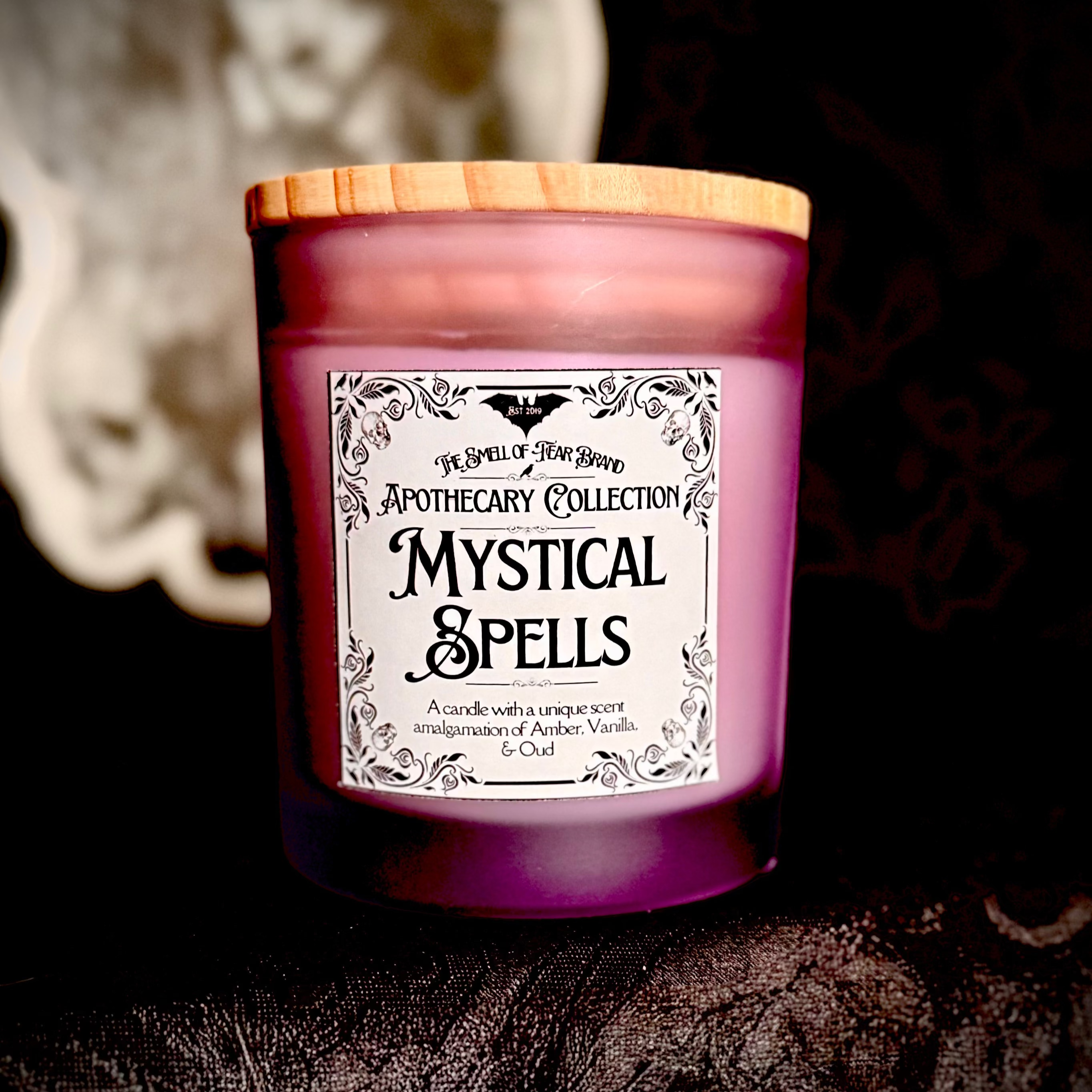 A purple candle labeled 'Mystical Moods' from the Apothecary Collection, with a wooden lid and decorative symbols in the background.