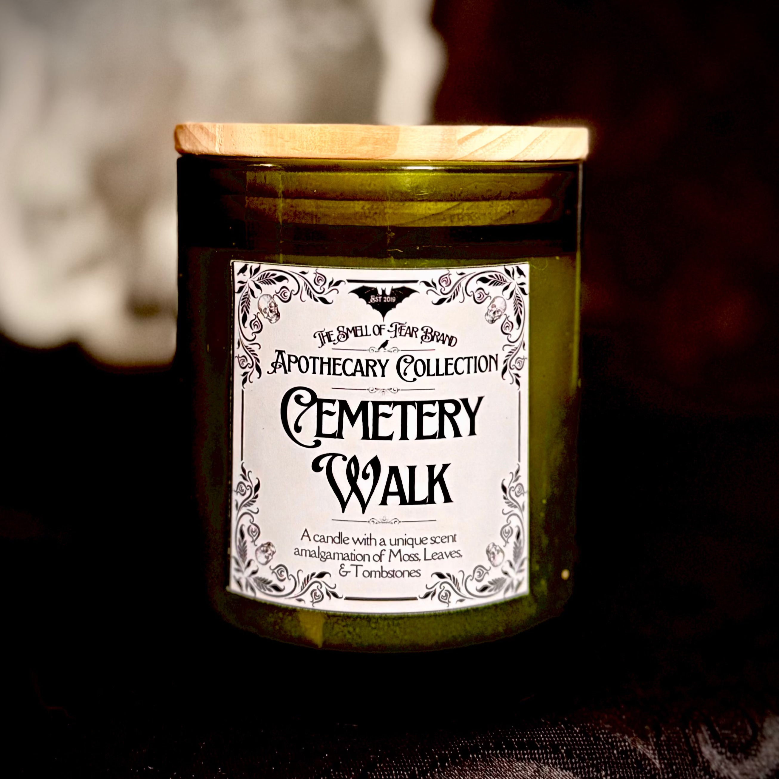 A green candle with a label reading 'Apothecary Collection: Cemetery Walk' placed on a grave in a cemetery.